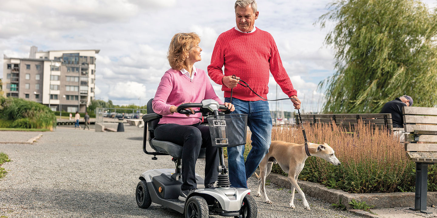 Promenadscooter med 4 hjul och liten korg fram. Kvinna åker på HS328 och man med hund går bredvid.